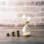 Jam pasir di samping tumpukan koin, menggambarkan konsep nilai waktu uang dalam keuangan